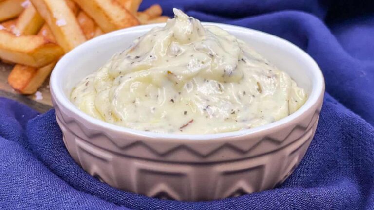 A bowl of creamy aioli sits on a blue cloth, with a portion of fries in the background.