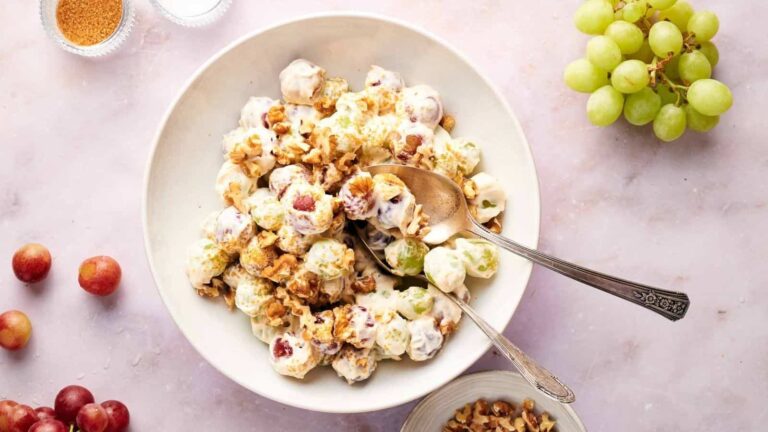 A bowl of creamy grape salad topped with chopped nuts on a light surface, surrounded by loose grapes and a small bowl of nuts.