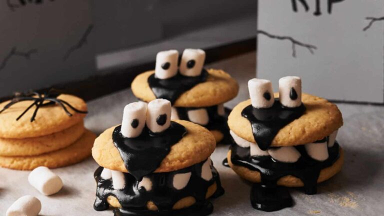 Three Halloween-themed cookies with black icing, marshmallow eyes, and teeth, resembling monsters. A stack of plain cookies is in the background.