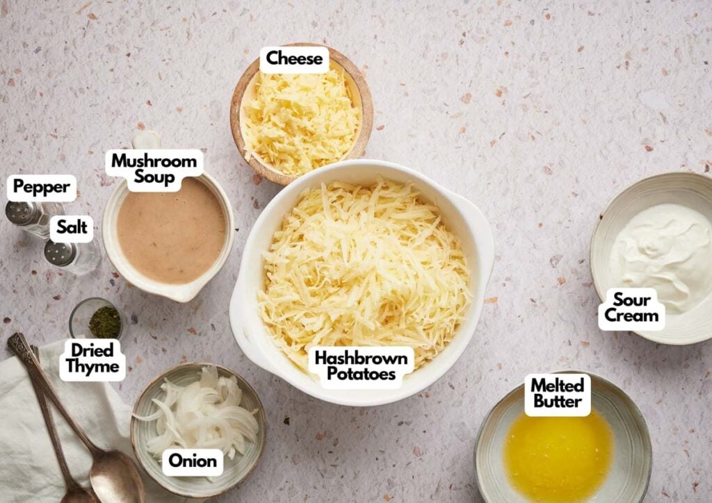 Top-down view of labeled ingredients for cowboy tater tot casserole, including hashbrown potatoes, cheese, mushroom soup, sour cream, melted butter, onion, dried thyme, salt, and pepper on a light surface.