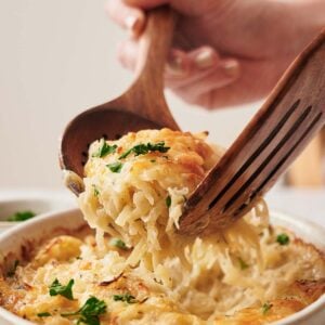 A hand serves cheesy baked pasta with wooden utensils from a ceramic dish, garnished with parsley.