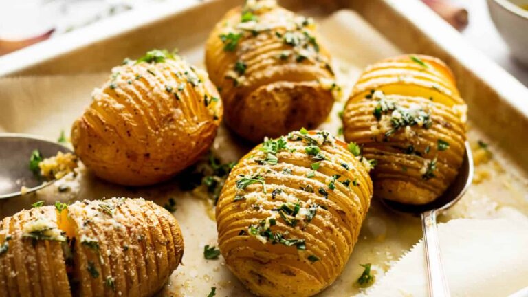 Baked hasselback potatoes topped with chopped herbs and grated cheese on a baking tray.