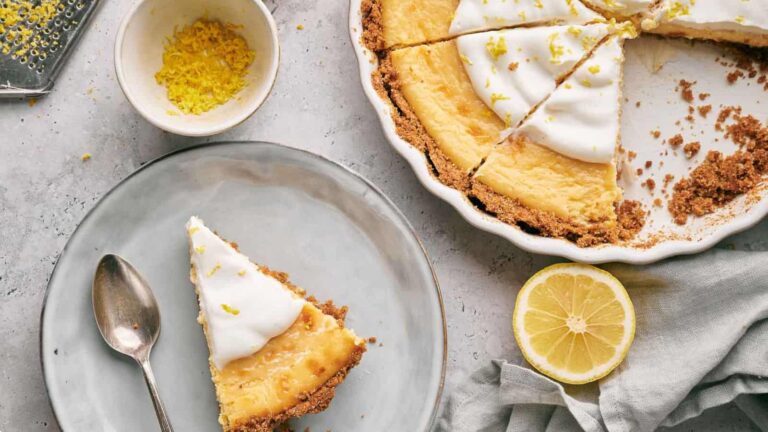 A lemon pie with a slice removed, topped with cream and lemon zest. A single slice on a plate with a spoon beside it. Additional lemon zest in a small bowl and half a lemon nearby.