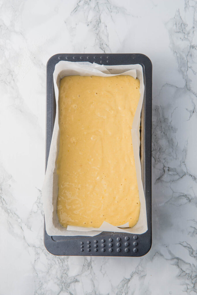 Lemon yogurt loaf batter in a parchment-lined rectangular baking pan on a marble countertop.