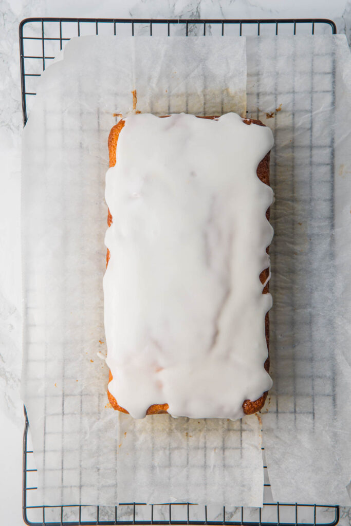 A lemon yogurt loaf cake covered with white icing sits on parchment paper on a cooling rack.