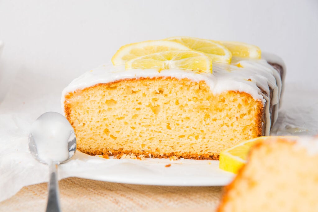 A lemon yogurt loaf with white icing is topped with lemon slices and sits on a white plate; a slice and a spoon are visible in the foreground.