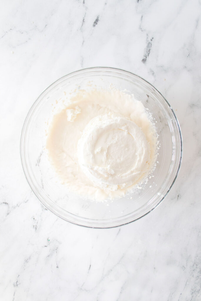 A clear glass bowl on a marble surface contains thick, creamy white cake batter reminiscent of Little Debbie Christmas Tree dip.