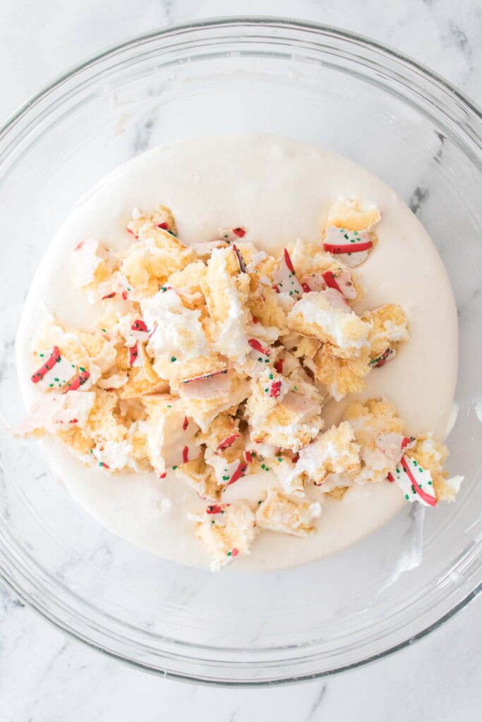 A glass bowl containing white icing and crumbled pieces of vanilla cake with sprinkles on a marble surface, reminiscent of Little Debbie Christmas Tree Dip.