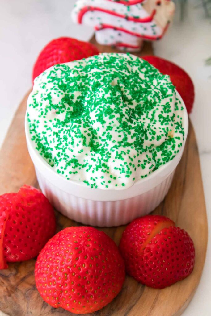 A bowl of creamy Little Debbie Christmas Tree dip topped with green sprinkles rests on a wooden board, surrounded by whole strawberries.
