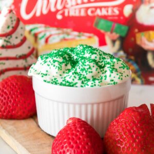 A white dish filled with Little Debbie Christmas Tree Dip, topped with whipped topping and green sprinkles, surrounded by strawberries on a wooden board. A festive Christmas-themed box is in the background.