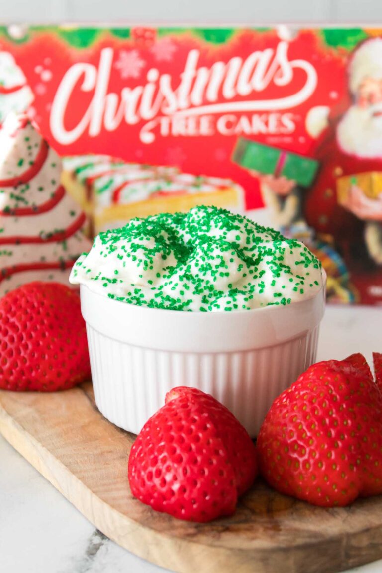 A white dish filled with Little Debbie Christmas Tree Dip, topped with whipped topping and green sprinkles, surrounded by strawberries on a wooden board. A festive Christmas-themed box is in the background.