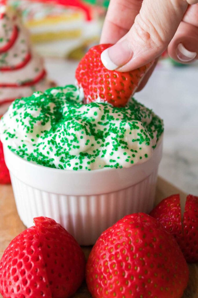 Hand dipping a strawberry into whipped cream, reminiscent of Little Debbie Christmas Tree Dip, topped with green sprinkles in a white ramekin, surrounded by more strawberries.