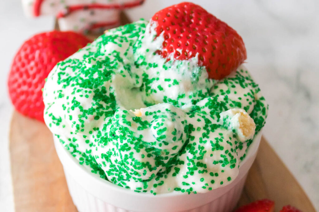 A small dish of creamy, Little Debbie Christmas Tree dip topped with green sprinkles and a whole strawberry, accompanied by additional strawberries in the background.