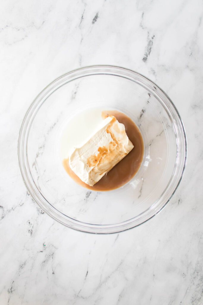 A glass bowl containing cream cheese, vanilla extract, and a liquid ingredient sits on a marble surface, perfect for creating a festive Little Debbie Christmas tree dip.