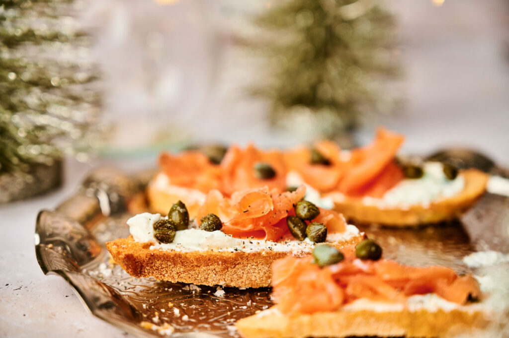 Crostini appetizers featuring slices of bread topped with cream cheese, smoked salmon, and capers are elegantly displayed on a silver platter, with blurred decorative trees in the background.