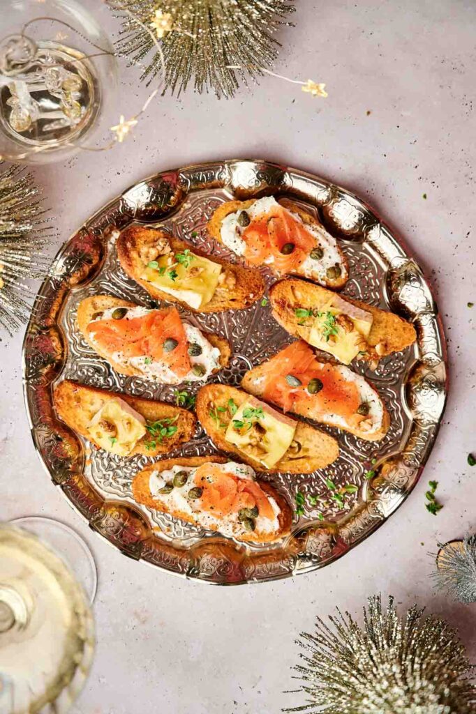 A silver platter with assorted crostini appetizers topped with smoked salmon, capers, herbs, and cheese. Surrounded by decorative elements and two glasses of white wine.
