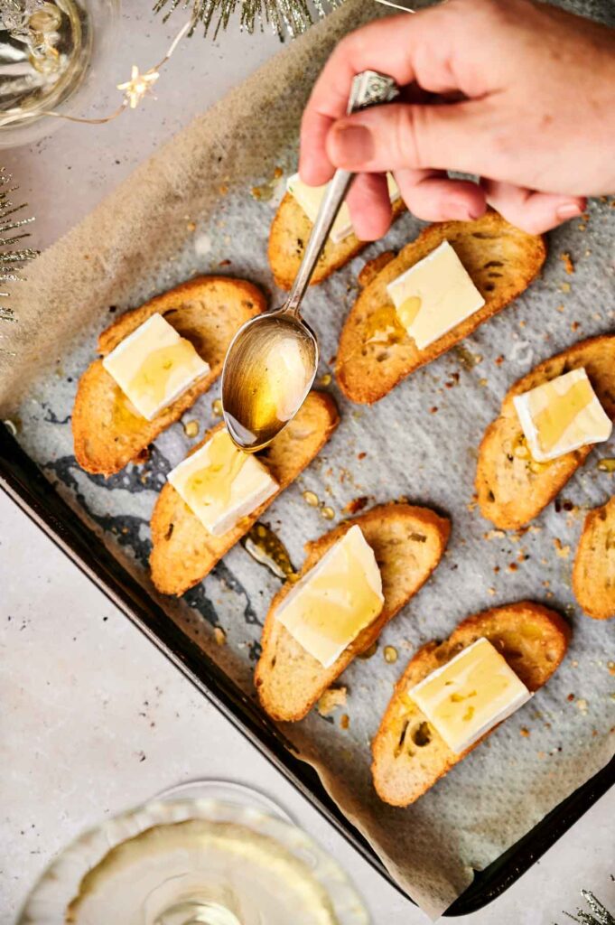 Hand drizzling honey over sliced bread topped with cheese on a baking tray, these crostini appetizers offer a delightful blend of sweet and savory.