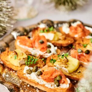A platter of crostini appetizers features toasted bread slices topped with cream cheese, smoked salmon, capers, and herbs, garnished with lemon wedges. Holiday-themed decorations are visible in the background.