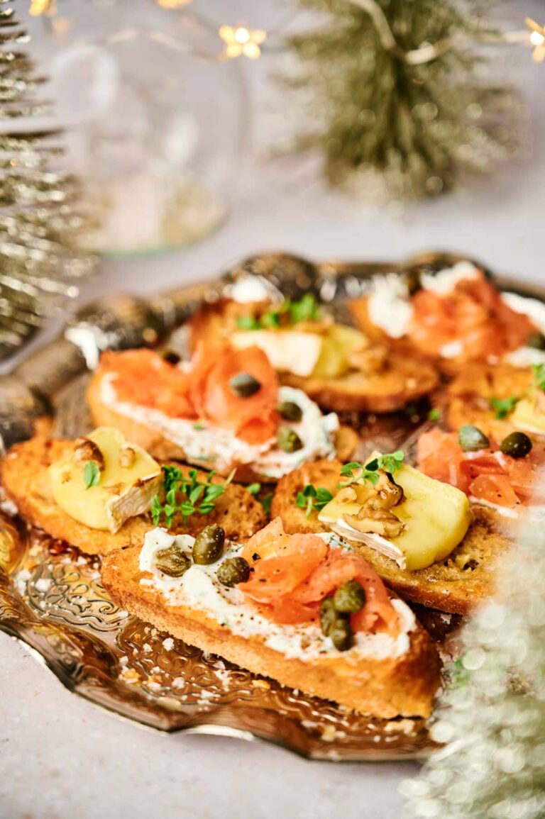 A platter of crostini appetizers features toasted bread slices topped with cream cheese, smoked salmon, capers, and herbs, garnished with lemon wedges. Holiday-themed decorations are visible in the background.