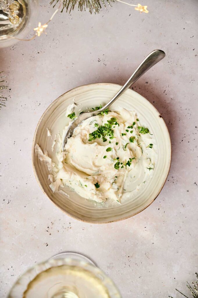 A bowl of creamy mashed potatoes topped with green herbs, accompanied by a spoon and set on a light beige surface. Crostini appetizers are artfully arranged nearby, while festive decorations add charm at the edges.