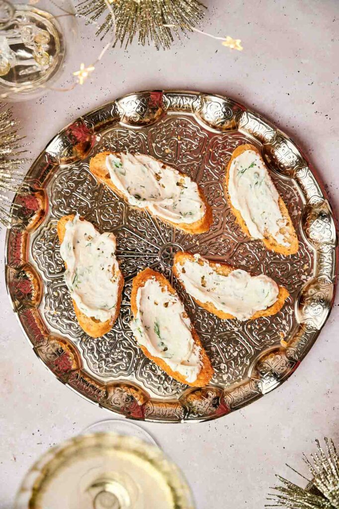 A silver tray showcases crostini appetizers, each featuring five slices of bread topped with a creamy white spread. Decorative elements enhance the scene, accompanied by a glass of white liquid nearby.