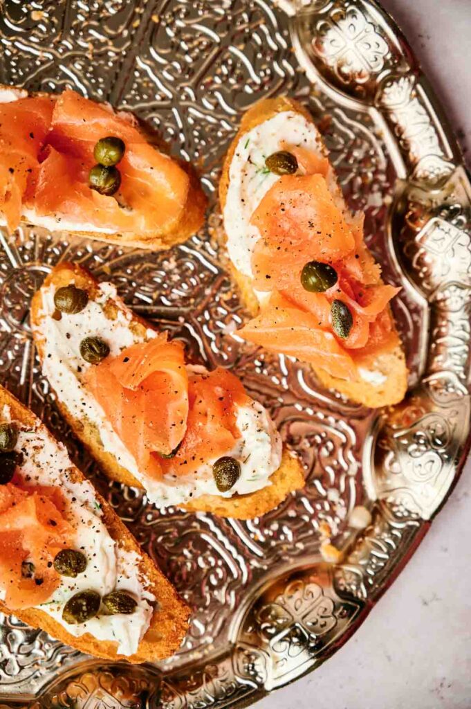 Four pieces of smoked salmon crostini with cream cheese and capers elegantly arranged on a decorative silver platter, perfect as appetizers.