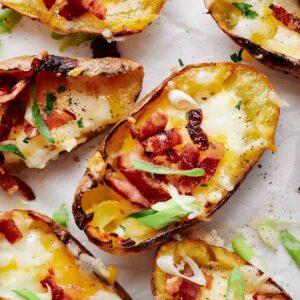 Loaded potato skins topped with melted cheese, crispy bacon bits, and sliced green onions rest on a parchment-lined surface.
