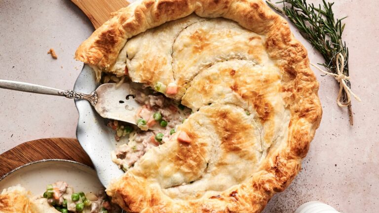 A freshly baked pie with a golden crust, a slice being served, revealing a filling of mixed vegetables and meat. A sprig of rosemary is nearby.