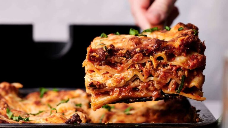 Close-up of a slice of lasagna being lifted from a baking dish. The lasagna has layers of pasta, cheese, and meat sauce, garnished with herbs.