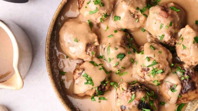 A bowl of meatballs covered in creamy sauce, garnished with chopped parsley, next to a small jug of additional sauce.
