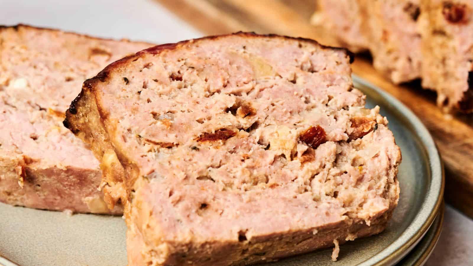 Close-up of several slices of meatloaf on a ceramic plate, showcasing the loaf's textured surface and browned edges with visible seasoning and ingredients.