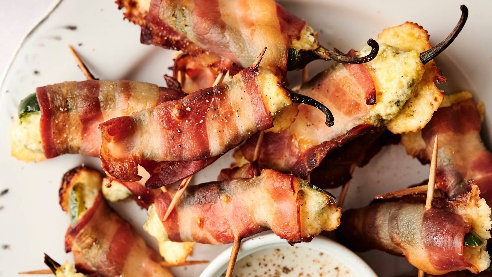 A plate of bacon-wrapped jalapeño poppers filled with cheese, secured with toothpicks, next to a small dish of dipping sauce.