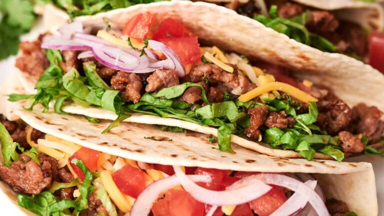 Close-up of tacos filled with seasoned meat, shredded cheese, chopped lettuce, diced tomatoes, and sliced red onions.