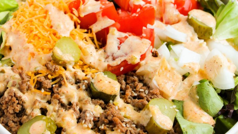 A salad featuring lettuce, ground beef, shredded cheese, sliced pickles, diced tomatoes, and onions, topped with a creamy dressing.