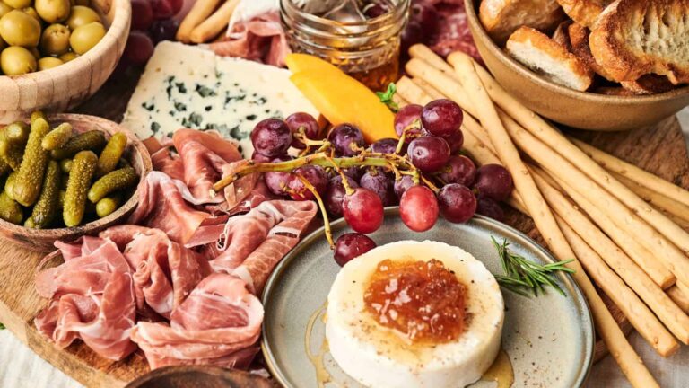 A charcuterie board with prosciutto, cheese, grapes, pickles, olives, breadsticks, and a small wheel of cheese topped with jam.