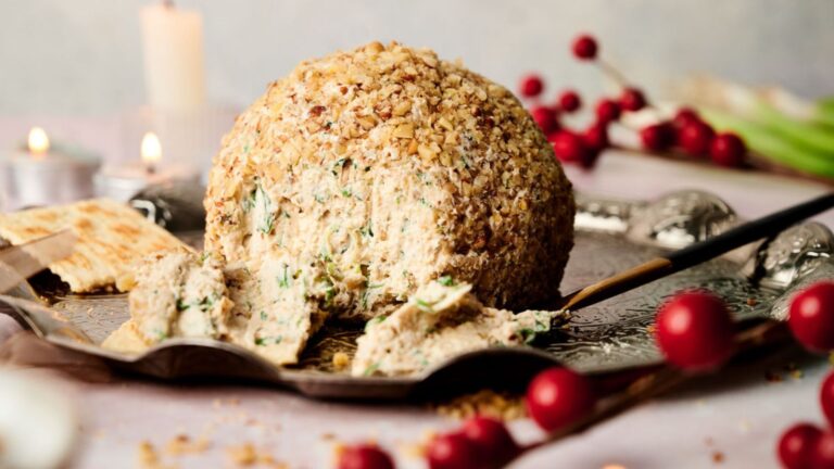 A round cheese ball coated in nuts is on a silver platter, partially spread on a cracker. Candles and red berries decorate the background.