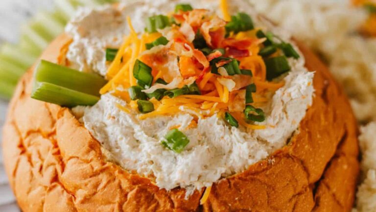 A bread bowl filled with creamy dip topped with shredded cheese, chopped green onions, and bacon bits, garnished with a celery stick.