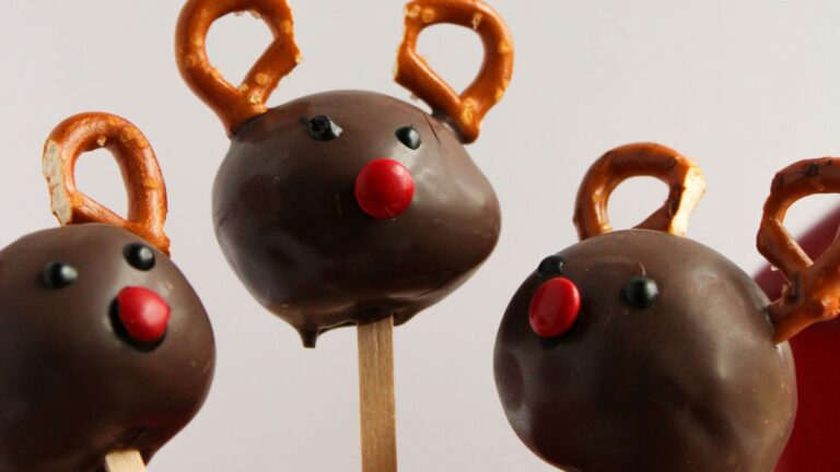 Three chocolate cake pops shaped like reindeer, with pretzel antlers and red candy noses, on wooden sticks.