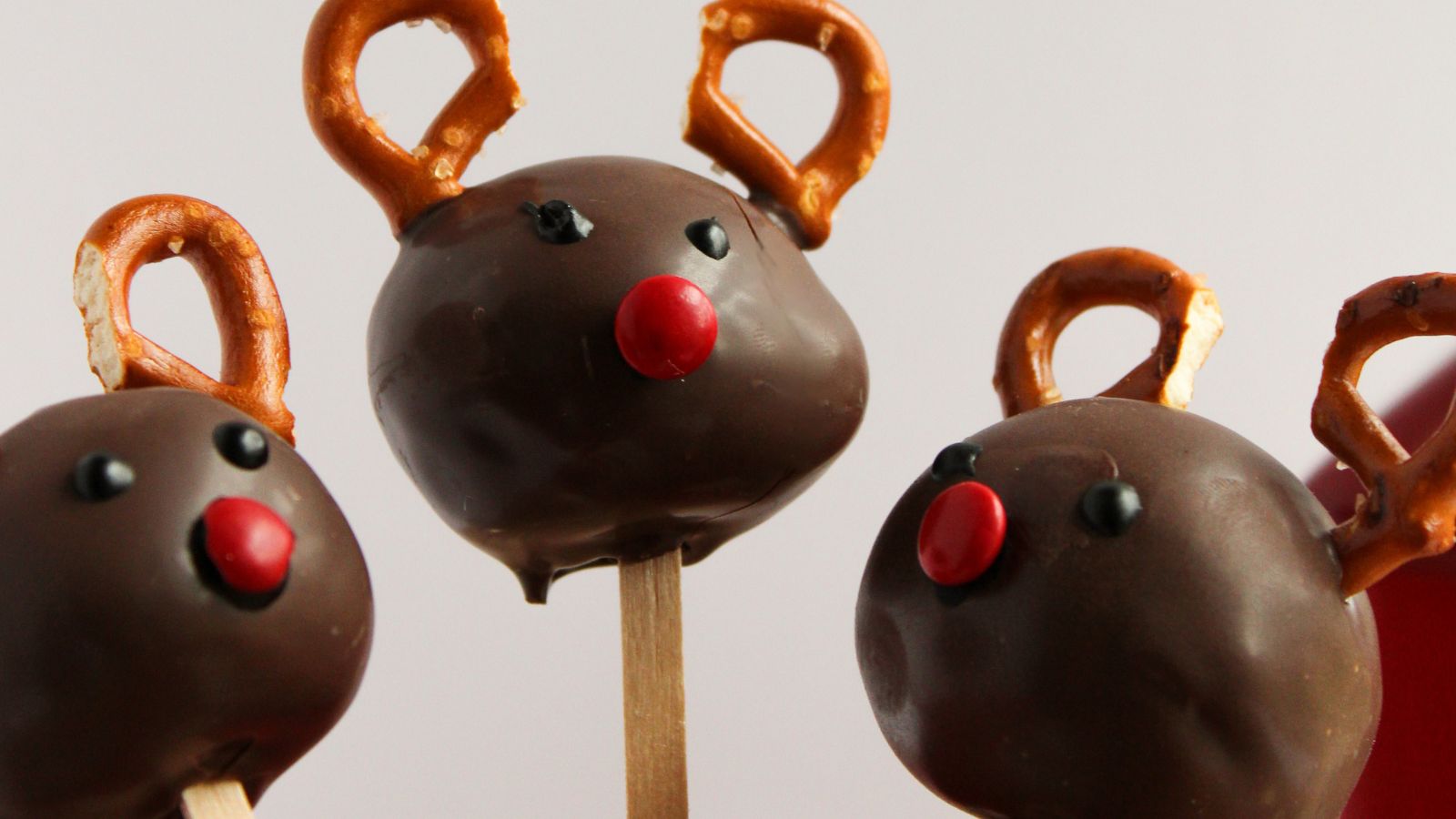 Three chocolate cake pops shaped like reindeer, with pretzel antlers and red candy noses, on wooden sticks.