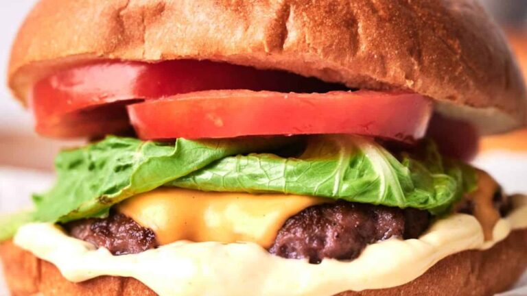 Close-up of a cheeseburger with a beef patty, lettuce, tomato slices, melted cheese, and sauce, inside a bun.