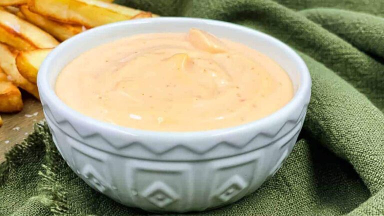 A white bowl filled with creamy orange sauce placed on a green cloth, with golden-brown fries visible in the background.