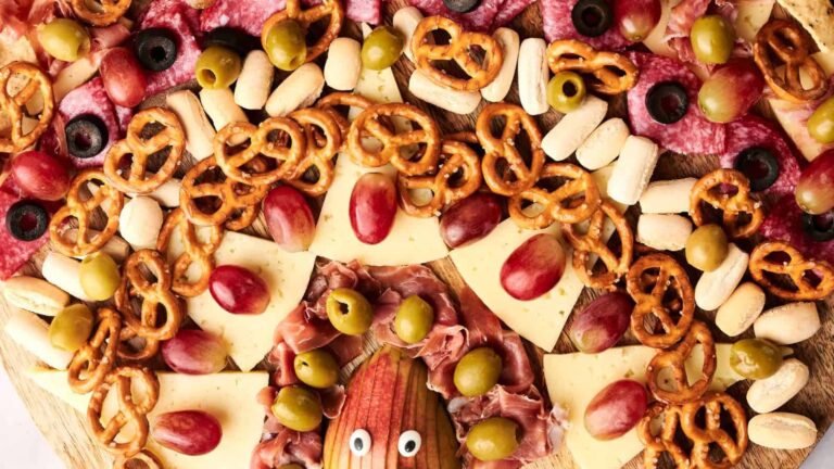 An arrangement of meats, cheeses, grapes, olives, pretzels, and breadsticks on a wooden surface, with an apple center featuring candy eyes.