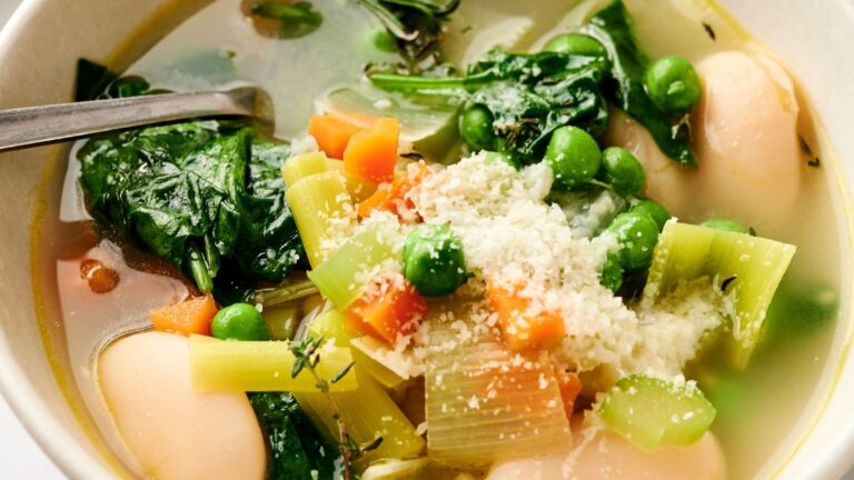 Bowl of vegetable soup with spinach, peas, carrots, leeks, and beans, topped with grated cheese, and a spoon on the side.