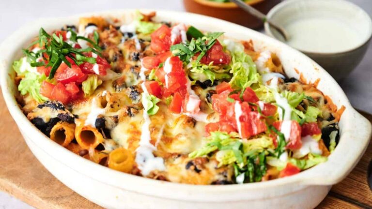 A baked pasta dish topped with melted cheese, diced tomatoes, lettuce, and a drizzle of sauce in a white casserole dish.