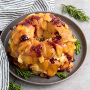 A bundt-shaped cranberry brie pull-apart bread topped with a glazed sauce and cranberries sits on a gray plate, garnished with rosemary sprigs. A striped cloth rests beside the plate on the gray surface.