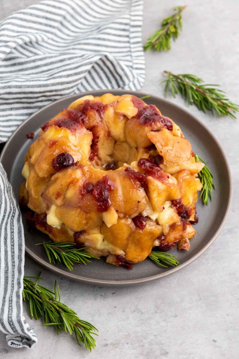 A bundt-shaped cranberry brie pull-apart bread topped with a glazed sauce and cranberries sits on a gray plate, garnished with rosemary sprigs. A striped cloth rests beside the plate on the gray surface.