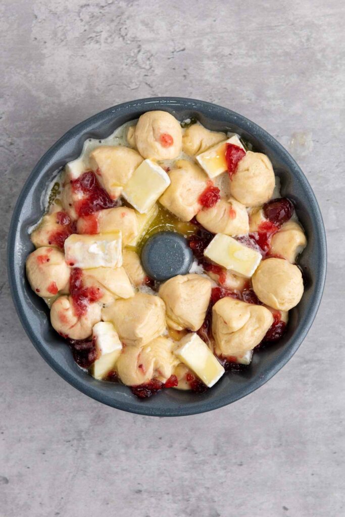Unbaked cranberry brie pull-apart bread in a bundt pan, adorned with chunks of creamy brie and dollops of raspberry jam on a gray background.