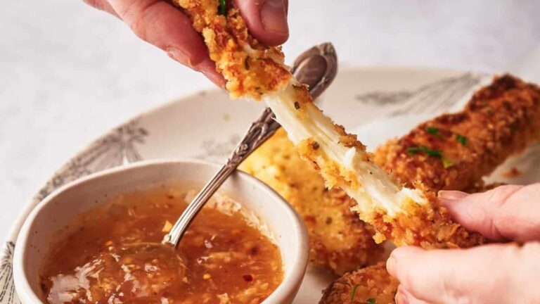 Hands dip a piece of mozzarella stick with melted cheese into a bowl of sauce on a plate.