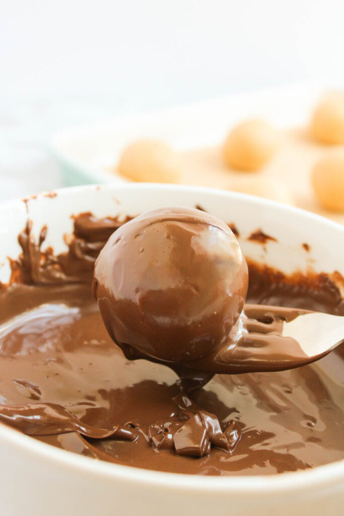 A spoon holds a round truffle being dipped in melted chocolate, reminiscent of decadent peanut butter balls, with several undipped truffles in the background.