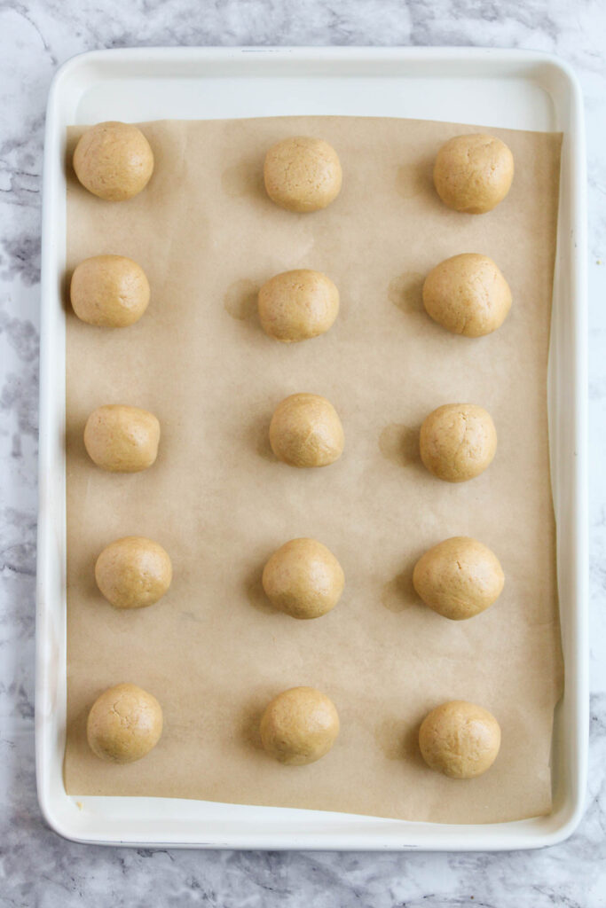 A baking tray lined with parchment paper holds 15 evenly spaced peanut butter dough balls, ready for baking.